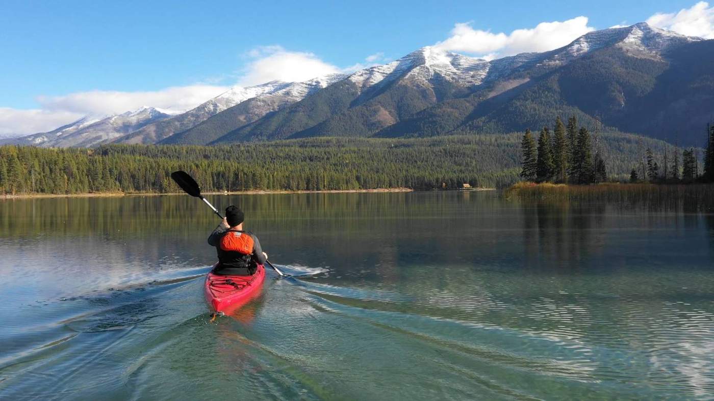 A kayaker in a red sea kayak paddles across a calm mountain lake toward snow-dusted, forested peaks
